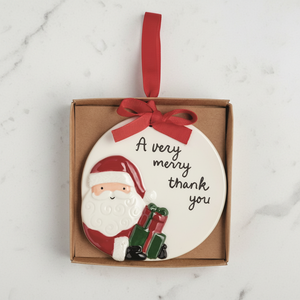 A round white ceramic Christmas ornament featuring a 3D Santa Claus figure and the text 'A very merry thank you', presented inside a brown gift box with a red ribbon loop.