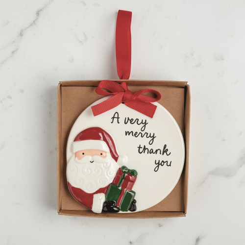 A round white ceramic Christmas ornament featuring a 3D Santa Claus figure and the text 'A very merry thank you', presented inside a brown gift box with a red ribbon loop.