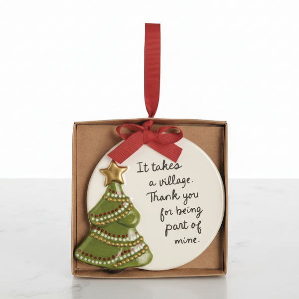A ceramic Christmas ornament in a square kraft gift box, featuring a white round ornament with the text 'It takes a village. Thank you for being part of mine.