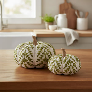 Two decorative fabric pumpkins with a cream base and olive green leafy vine pattern, featuring natural wood stems, displayed on a light gray surface against a white background.