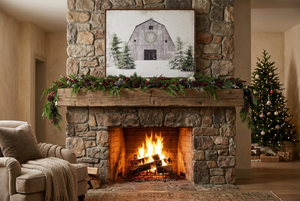 A white barn and pine tree print hanging above a stone fireplace with a crackling fire, a wood mantel draped in greenery, and a cozy armchair nearby.
