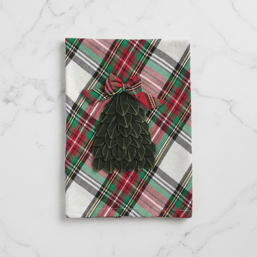 A rectangular kitchen towel with a red, green, and white plaid pattern, featuring a dark green tiered Christmas tree applique tied with a matching plaid bow at the top.