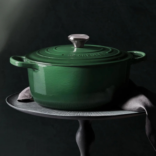 A Artichaut Le Creuset enameled cast iron Dutch oven with a matching lid, displayed on a dark metal pedestal against a dark background.
