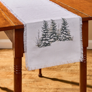 White woven cotton table runner with fringed edges and a watercolor of three snow-covered spruce trees, draped over a wooden table.