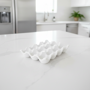A white ceramic egg-shaped tray with wavy edges sits on a white marble kitchen island, with a softly blurred modern kitchen in the background.