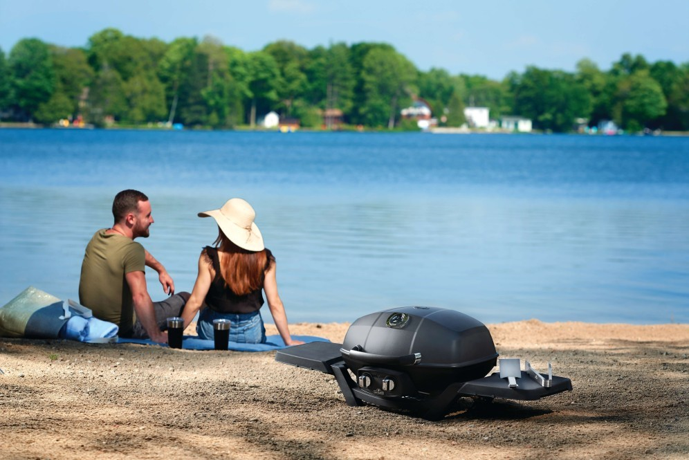 Two people sitting by a lake with a portable grill on the sand.