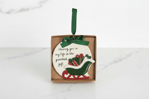 A round ceramic Christmas ornament in a brown gift box, featuring a white background with the phrase 'Having you in my life is the greatest gift' in black script, a green and red sleigh, and a green hanging ribbon.