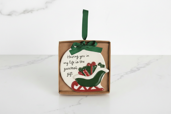 A round ceramic Christmas ornament in a brown gift box, featuring a white background with the phrase 'Having you in my life is the greatest gift' in black script, a green and red sleigh, and a green hanging ribbon.
