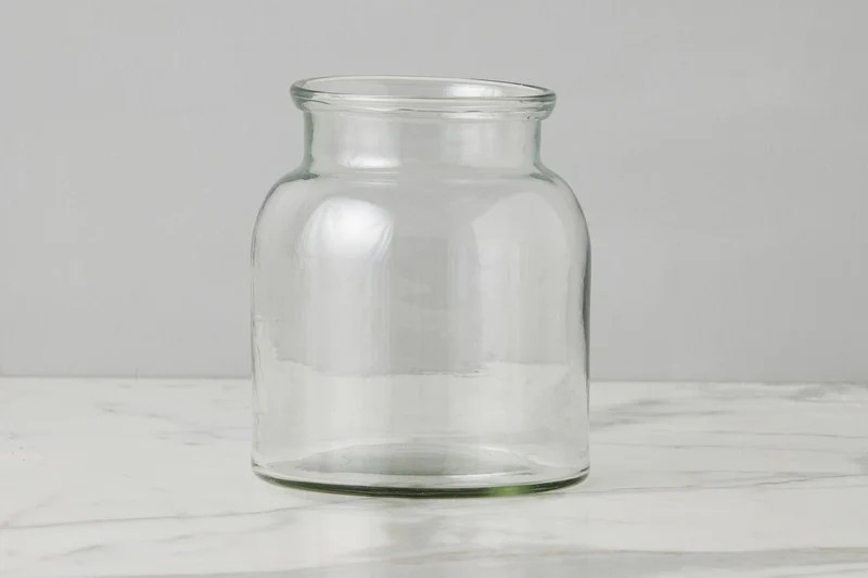 A clear glass jar with a wide mouth and rounded body, standing on a light marble surface against a neutral gray background.