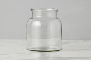 A clear glass jar with a wide mouth and rounded body, standing on a light marble surface against a neutral gray background.