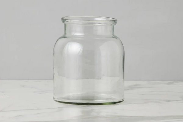 A clear glass jar with a wide mouth and rounded body, standing on a light marble surface against a neutral gray background.