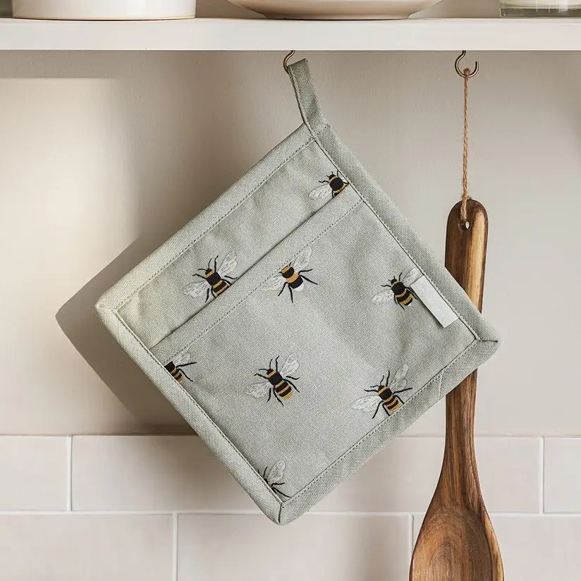 A square kitchen pot holder with  bumblebees, hanging from a metal hook on a kitchen shelf next to a wooden spoon.