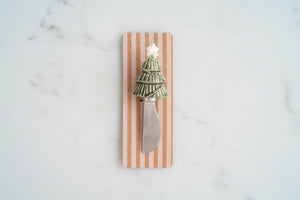 A decorative cheese knife with a miniature green Christmas tree handle and a silver blade, mounted on a vertical wooden plaque with pink and white stripes.