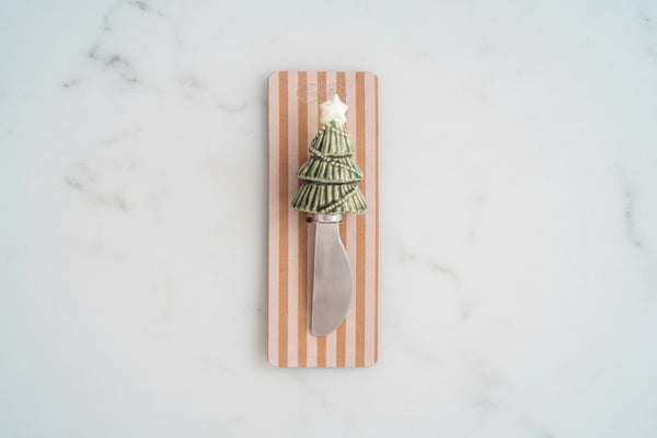 A decorative cheese knife with a miniature green Christmas tree handle and a silver blade, mounted on a vertical wooden plaque with pink and white stripes.