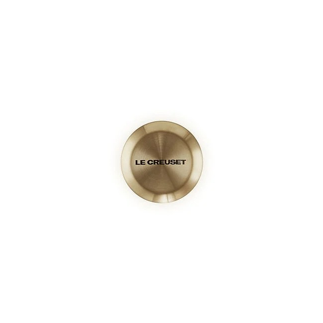 A single round Le creuset knob in a light gold finish, featuring the 'LE CREUSET' brand name printed on its top surface, set against a plain white background.
