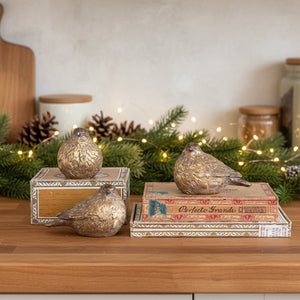 Three metallic gold bird figurines with textured feathers, styled on vintage books and a small decorative box against a light gray background.