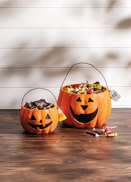 Two carved jack-o'-lantern candy baskets filled with Halloween treats sit on a dark wood floor against a white paneled wall with soft shadows.